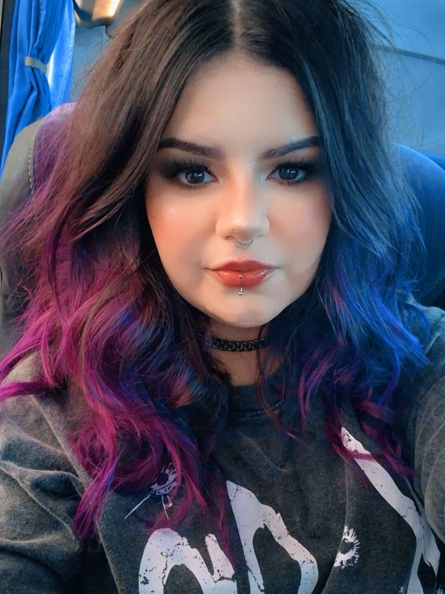Woman with medium black wavy hair with purple tips, fair skin, dark brown eyes. She wears dark makeup, red lip gloss, a septum nose ring, and a lower lip piercing. She is wearing a patterned grey t-shirt and a silver choker. The background shows the blurred interior of a bus.